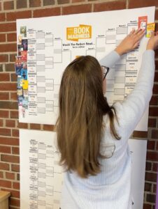 Librarian moves tiny book covers across a banner to show how the book is faring in a tournament-format contest that allows kids to vote for their favorite book. 